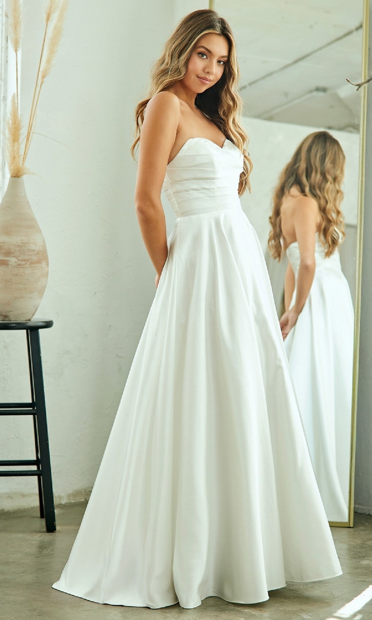 A woman indoors stands by a mirror and vase with pampas grass, wearing the Amelia Couture 5076 Classic Simple White Prom Dress in strapless satin.